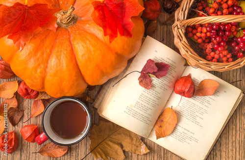 Autumn still life; open book, mug of tea; orange squash; basket with red berries and dry leaves on a wooden background top view