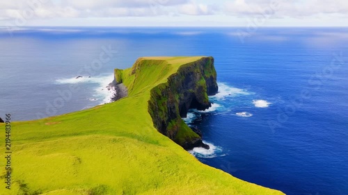 Grassy coastal cliff meets the open ocean as waves break along a scenic headland beneath a partly cloudy. Travel promotion, tourism advertising, and nature documentaries.