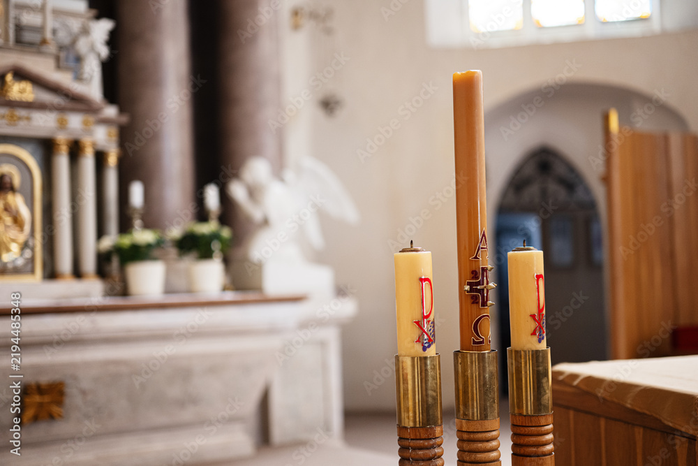 Fototapeta premium Three decorated altar candles with Paschal symbols in a church interior near an angel statue and altar during Easter liturgy