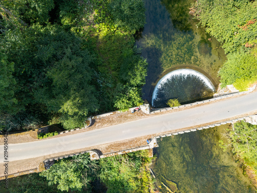 Aerial view of the River Teme flowing under a stone bridge with a semicircular weir surrounded by lush green trees in Ludlow, Shropshire, United Kingdom.