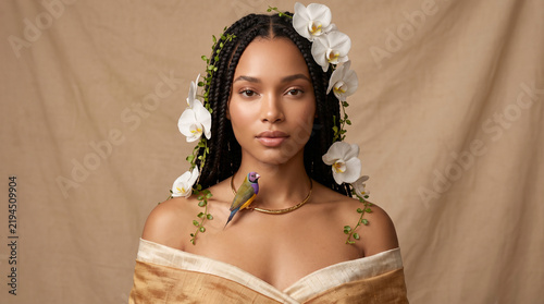 Serene conceptual beauty portrait of a beautiful young Black woman adorned with white orchids, green vines, and a small colorful bird resting on her chest against a draped fabric backdrop.