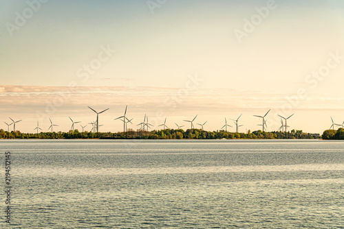 Wind farm with multiple wind turbines along a green shoreline viewed across calm water. Renewable energy concept.