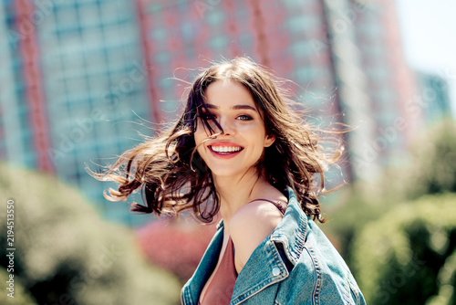 Smiling young woman in denim jacket enjoying a windy sunny day in the city, candid outdoor portrait conveying happiness and carefree summer energy
