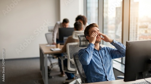 Exhausted employee rubbing eyes at office desk, tired male employee suffering from eye strain and headache, stressed worker feeling burnout in modern workspace