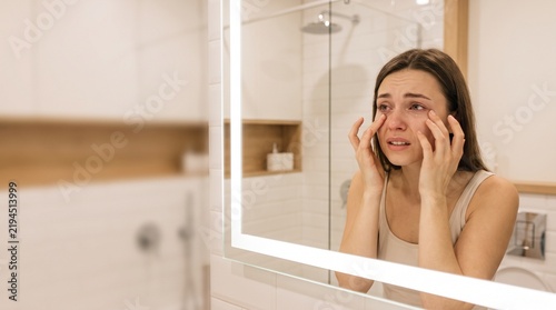 Crying woman looking in bathroom mirror, upset young female with red eyes and tears, emotional distress and mental health concept in modern interior