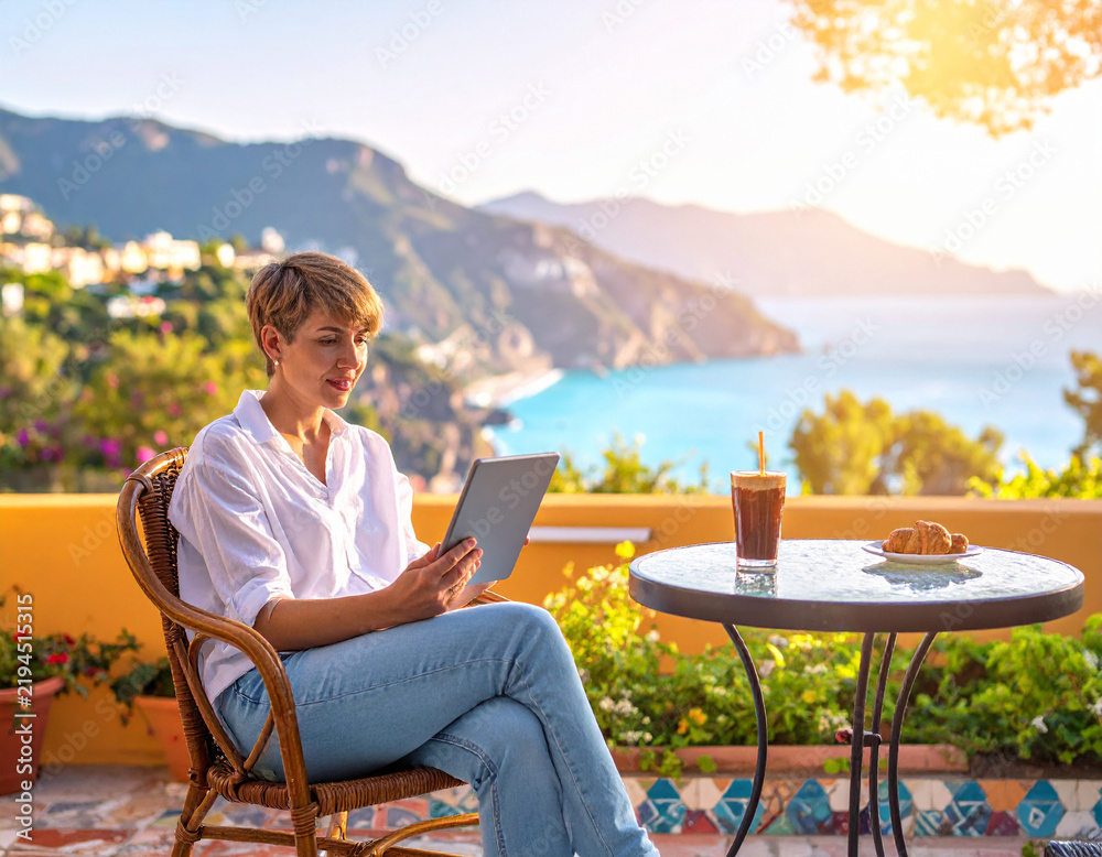 Fototapeta premium A serene woman works on a digital tablet at a Mediterranean terrace cafe during sunset. This peaceful scene evokes freedom and luxury travel, perfect for digital nomad or lifestyle marketing.