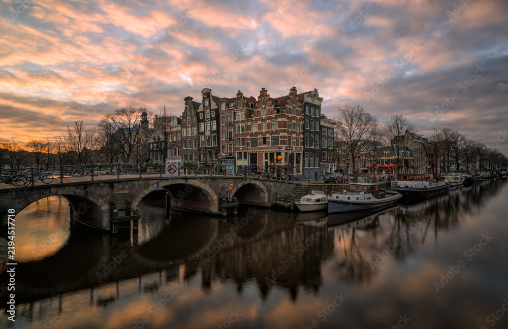 Obraz premium Amsterdam canal at sunset with historic bridge and houseboats reflecting in calm water