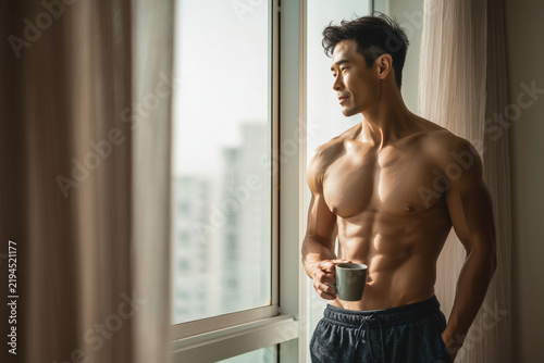 Shirtless Asian Man Holding Coffee Mug By Large Window In Morning Light At Home
