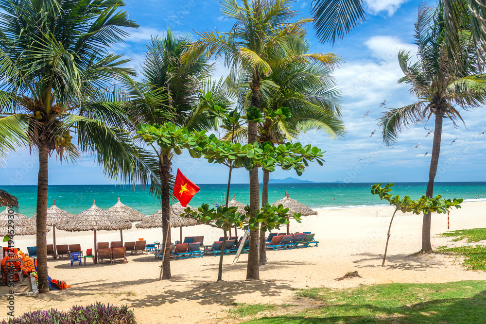 Fototapeta premium My Khe beach in Da Nang with palm trees, Vietnamese flag, thatched umbrellas, sun loungers, and birds flying over turquoise sea