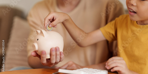 Asian boy putting coin in piggy bank with parent for financial literacy and savings habit