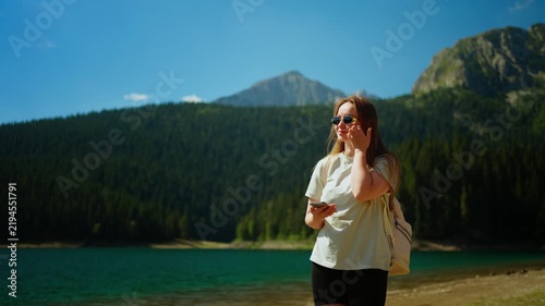 Young woman walking along a tranquil mountain lake shoreline, enjoying sunny weather, forest scenery, and peaceful nature during an outdoor summer getaway hike