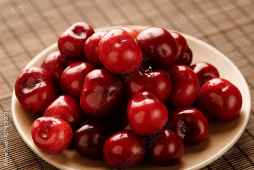 Close-up of glossy, ripe red cherries piled on cream ceramic plate over bamboo mat, emphasizing vibrant color, smooth texture and natural freshness for food, recipe, healthy lifestyle seasonal themes