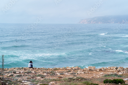 Foto A solitary woman sits on a rocky coastline watching turquoise waves and a misty horizon, evoking solitude, reflection and a peaceful, contemplative mood in a natural seaside landscape