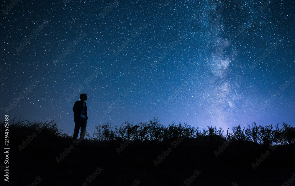 Fototapeta premium A person's silhouette stands on a hilltop, gazing at the vast starry night sky with the milky way stretching across the blue horizon, surrounded by dark vegetation.