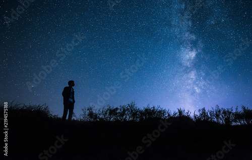 A person's silhouette stands on a hilltop, gazing at the vast starry night sky with the milky way stretching across the blue horizon, surrounded by dark vegetation.