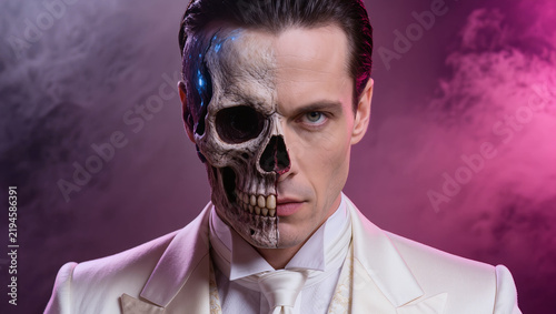 Elegant young man in a white tuxedo with slicked-back platinum-blonde hair, featuring dramatic decaying skull special-effects makeup and vivid pink, purple, crimson, and blue tones.