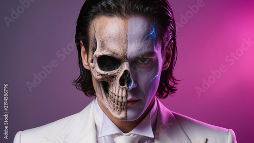 Elegant young man in a white tuxedo with slicked-back platinum-blonde hair, featuring dramatic decaying skull special-effects makeup and vivid pink, purple, crimson, and blue tones.