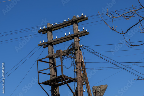 A double wooden utility pole with white insulators stands against a vibrant blue sky, highlighting infrastructure designed for electrical power distribution in an industrial area