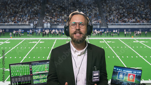 White Male Broadcaster Wearing Glasses, Headset Microphone and Press Pass Speaks to Camera While Covering a Packed Football Arena, Dual Laptops Displaying Game Footage and Club Emblems.
