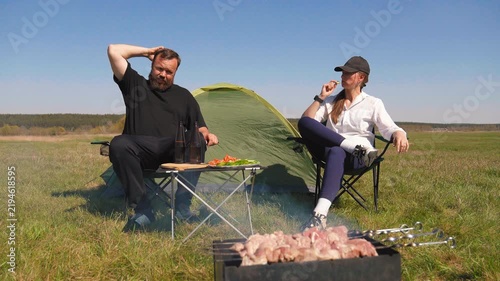 Bearded man and young woman relaxing and talking near a tent in a grassy field, enjoying a barbecue picnic with fresh vegetables and drinks on a folding table during their camping trip