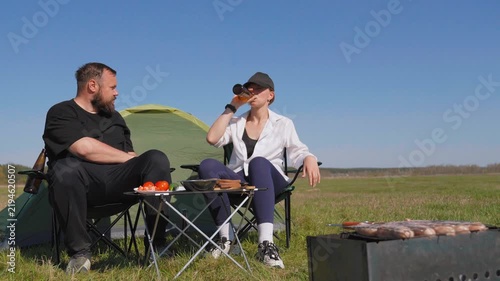 Young man and woman sitting on camping chairs in nature, drinking beer from bottles and enjoying a relaxing picnic with a tent set up in a beautiful green meadow under a clear sunny sky