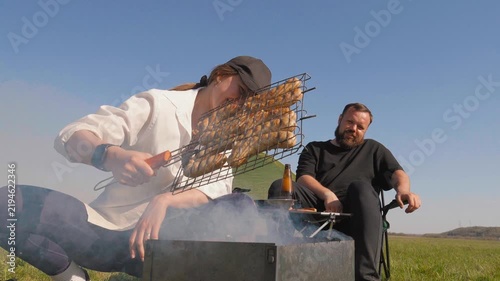 Smiling young woman grilling chicken and sausages on a portable charcoal grill while her bearded boyfriend relaxes in a chair, enjoying a sunny day of camping and outdoor picnicking