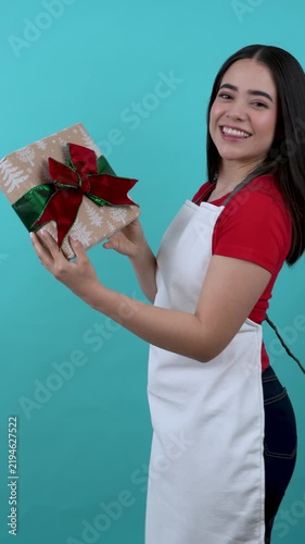 Young latina female shop assistant in an apron holding a festive christmas present on a teal background