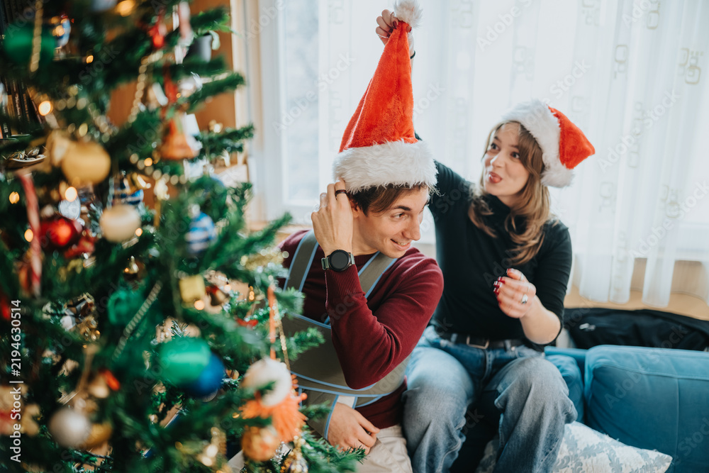 Naklejka premium A young couple wearing Santa hats sit by a decorated Christmas tree and laugh while they decorate. The pair share a playful moment trimming ornaments and enjoying a cozy holiday scene.