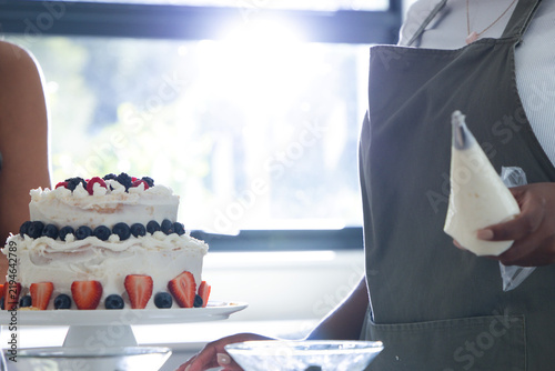 Two-tier round cake is sitting on white pedestal near bright window, topped with assorted berries
