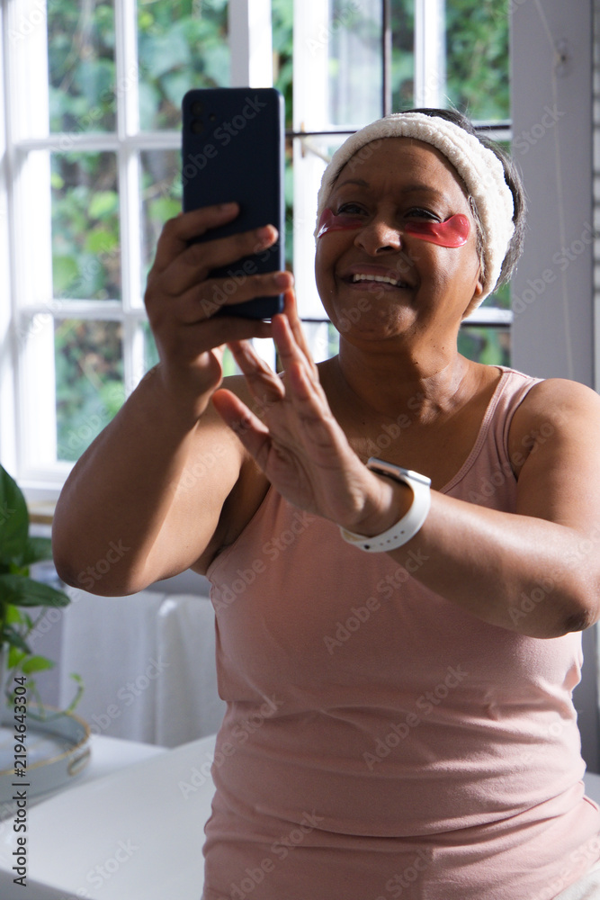 Obraz premium Mature African woman holding smartphone, smiling, taking selfie in bathroom with under-eye patches