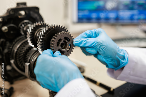 Close-up of a technician in blue gloves inspecting metal gears in an engine assembly, with a computer screen blurred in the background.
