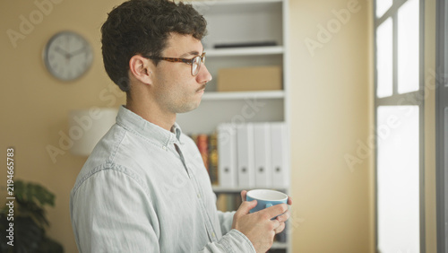 Quadro su tela Man holding blue mug and looking out window in building office, wearing glasses and casual shirt while pausing midbreak; quiet contemplation