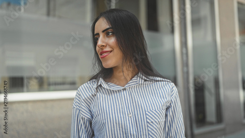 Фотография Young woman with visible face and slight smile, head turned to right looking sideways at storefront building; quiet confidence