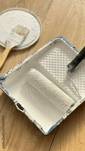 Painting Supplies with Roller and Brushes on Wooden Floor. Paint Roller and Brush with White Paint on Wooden Floor. Painting Tools and Supplies on Wooden Floor