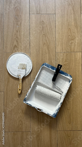Painting Supplies with Roller and Brushes on Wooden Floor. Paint Roller and Brush with White Paint on Wooden Floor. Painting Tools and Supplies on Wooden Floor