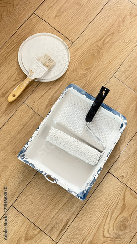 Painting Supplies with Roller and Brushes on Wooden Floor. Paint Roller and Brush with White Paint on Wooden Floor. Painting Tools and Supplies on Wooden Floor