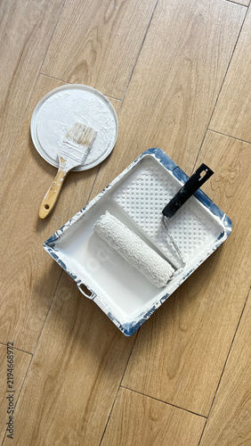 Painting Supplies with Roller and Brushes on Wooden Floor. Paint Roller and Brush with White Paint on Wooden Floor. Painting Tools and Supplies on Wooden Floor
