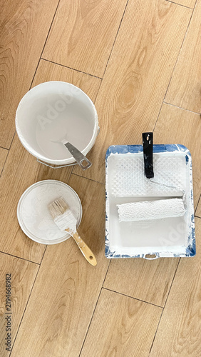 Painting Supplies with Roller and Brushes on Wooden Floor. Paint Roller and Brush with White Paint on Wooden Floor. Painting Tools and Supplies on Wooden Floor