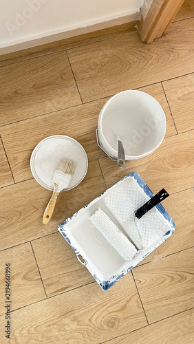 Painting Supplies with Roller and Brushes on Wooden Floor. Paint Roller and Brush with White Paint on Wooden Floor. Painting Tools and Supplies on Wooden Floor