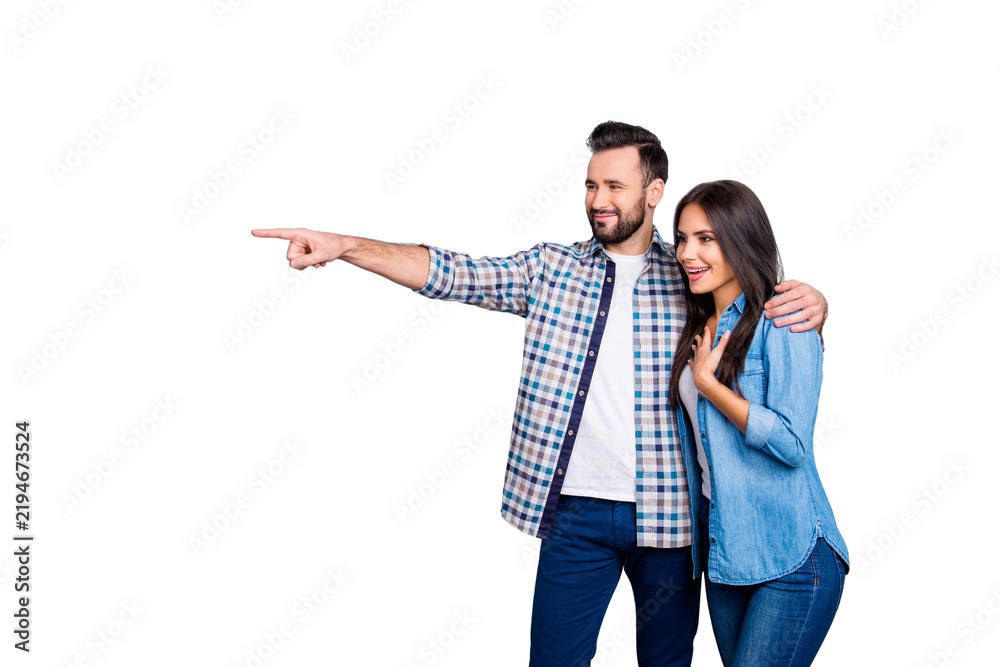 Fototapeta premium Bearded, smiling man embracing his excited, charming girlfriend and showing to her something with forefinger over grey background