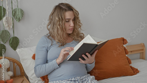 Obraz na płótnie Pregnant woman reading a book, hand turning a page while seated on a bed in a bedroom; quiet reflection calm