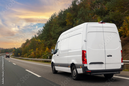 White courier delivery van drives highway autumn forest dramatic warm sunset sky. Logistics, transport and delivery concept blends motion, travel and seasonal landscape in calm rural scene