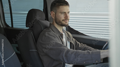 Bild auf Leinwand Man driving with hands on steering wheel inside car beside corrugated metal building facade, gazing forward in a steady pose; calm contemplation