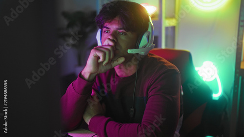 Fotografie Man wearing headset clasps hands under chin at gaming studio desk with keyboard, mouse and neon controller sign; quiet contemplation