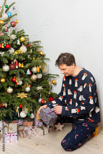 A 53-year-old man kneels by a Christmas tree in his home on Christmas Day. He examines a gift carefully as holiday decorations surround him. The moment is filled with family joy and cheer