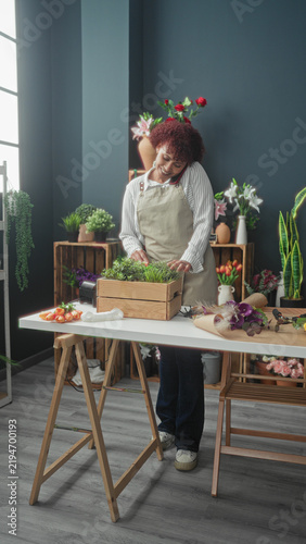 Konstfotografi Woman florist arranges plants in a wooden crate, hands trimming greenery while holding smartphone at ear in a studio; calm care