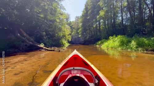 POV kayaking along a shallow forest river on a sunny summer day, red kayak moving through calm clear water surrounded by lush green woodland.