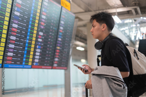 Young traveler checking a flight information board at an airport while holding a smartphone, wearing headphones and carrying a backpack, representing travel planning, departure, adventure, and journey