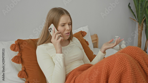 Картината върху платно Young woman on bed holds smartphone to ear, hand palm up gesture and furrowed face in a building; frustration complaint