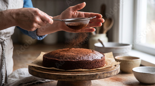 Person dusting a freshly baked chocolate cake with cocoa powder using a fine mesh sieve